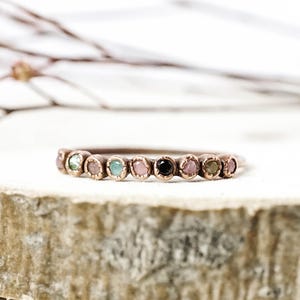May include: A delicate copper band ring with a row of colorful gemstones. The stones are set in a bezel setting and are arranged in a rainbow pattern. The ring is a beautiful and unique piece of jewelry.