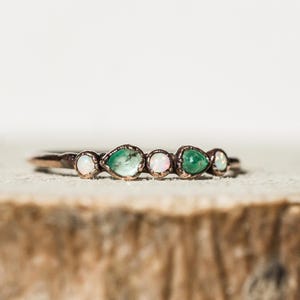Può includere: Un anello delicato con una fascia color rame, con cinque pietre preziose. Le pietre preziose alternano tra opali bianchi ovali e smeraldi verdi a forma di pera. L'anello è posto su uno sfondo neutro.