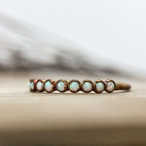 May include: A delicate ring with a bronze band, set with a series of small, round opals. The opals are a milky white color, and the ring is set against a blurred background.