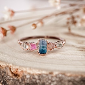 Può includere: Un anello in oro rosa con una montatura di pietre preziose blu, rosa e bianche. L'anello è su una superficie di legno.