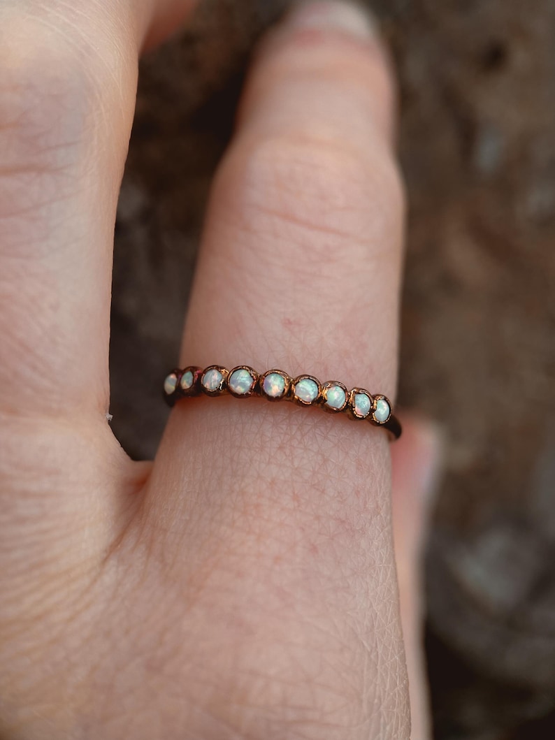May include: A delicate ring featuring a row of small, round opals set in a thin, dark metal band. The opals have a milky white color and are evenly spaced around the ring. The ring is shown on a finger.