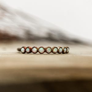 May include: A delicate ring featuring a series of small, round, opalescent stones set in a bronze-colored band. The ring is displayed on a light brown surface, showcasing its subtle sparkle and elegant design. A piece of jewelry.