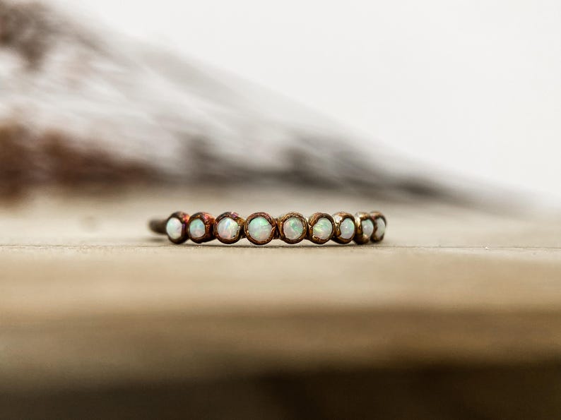 May include: A delicate ring featuring a series of small, round, opalescent stones set in a bronze-colored band. The ring is displayed on a light brown surface, showcasing its subtle sparkle and elegant design. A piece of jewelry.