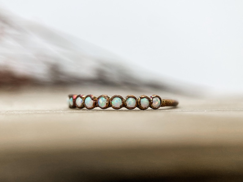 May include: A delicate ring with a bronze band, set with a series of small, round opals. The opals are a milky white color, and the ring is set against a blurred background.