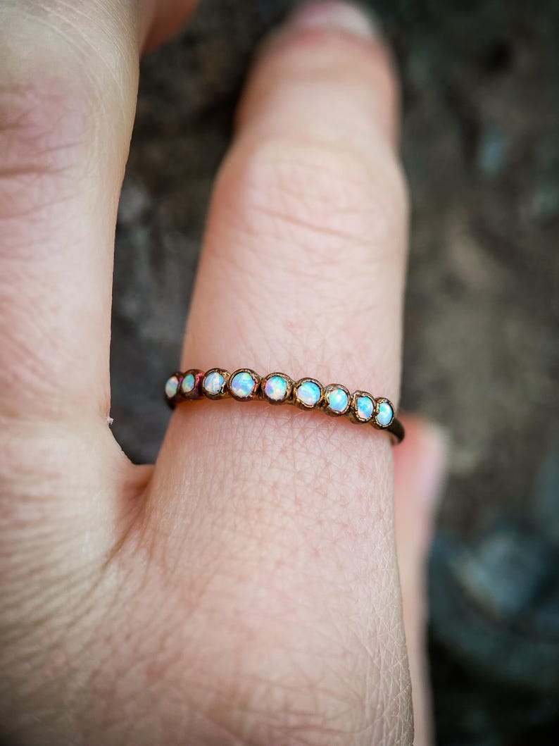 May include: A delicate ring featuring a row of small, round opals set in a thin, bronze-colored band. The opals display a range of iridescent colors, including white, blue, and hints of pink. The ring is shown on a finger.