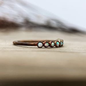 May include: A delicate ring with a copper-colored band. The ring is adorned with six small, round, opalescent stones. The stones have a subtle iridescence, reflecting shades of white, blue, and green. The ring is set against a blurred background.