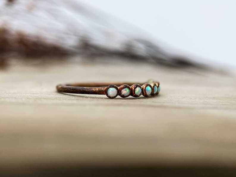 May include: A delicate ring with a copper-colored band. The ring is adorned with six small, round, opalescent stones. The stones have a subtle iridescence, reflecting shades of white, blue, and green. The ring is set against a blurred background.