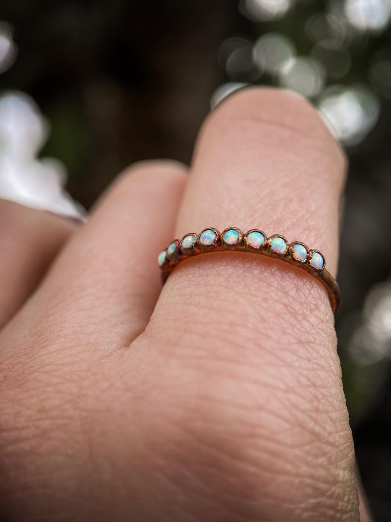 May include: A delicate gold ring adorned with small, iridescent opals. The ring features a thin band and a series of opals set closely together, creating a subtle yet eye-catching design. The opals display a range of colors, adding a touch of elegance.