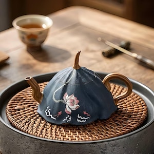 May include: A navy blue ceramic teapot with a lotus flower design, set on a woven mat within a dark gray tray. The teapot has a curved handle and spout, and a small cup of tea is in the background.