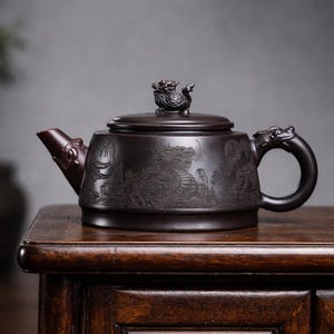 Peut inclure: Théière en céramique marron foncé avec des gravures en relief de dragons et de vagues. Le couvercle est orné d'une petite figurine de dragon. La poignée et le bec présentent également des motifs de dragon. La théière est posée sur une surface en bois foncé.