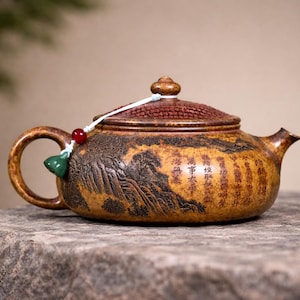 May include: A decorative brown teapot with a textured surface and a lid. The teapot features a handle, spout, and a small decorative bead. The body of the teapot has a landscape design and text. The teapot is sitting on a stone surface.