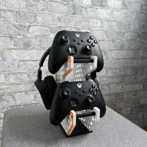 Könnte beinhalten: Ein schwarzer Gaming-Controller-Ständer mit zwei Controllern und einem Headset. Der Ständer ist weiß mit orangefarbenen Akzenten und hält die Controller und das Headset an Ort und Stelle. Die Controller sind schwarz mit farbigen Knöpfen.
