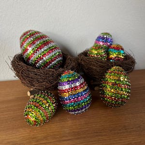 Könnte beinhalten: Dekorative Ostereier, die mit bunten Pailletten bedeckt sind. Einige Eier liegen in kleinen Zweignestern. Die Eier weisen eine Vielzahl von Farben auf, darunter Rot, Grün, Silber und Regenbogenfarben, was eine festliche Präsentation ergibt.