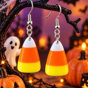 May include: A pair of dangle earrings shaped like candy corn, featuring a smooth, tri-colored design with a white top, orange middle, and yellow base. The earrings are attached to silver-toned metal fishhook wires with a small jump ring connector.