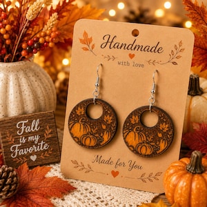 May include: A pair of round wooden drop earrings featuring laser-engraved pumpkin and autumn leaf designs. The earrings are attached to a tan cardboard display card printed with the text Handmade with love and Made for You in black ink.