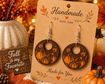 Pode incluir: Um par de brincos pendentes redondos de madeira com desenhos de abóboras e folhas de outono gravados a laser. Os brincos estão presos a um cartão de papelão bege com o texto Handmade with love e Made for You impresso em tinta preta.