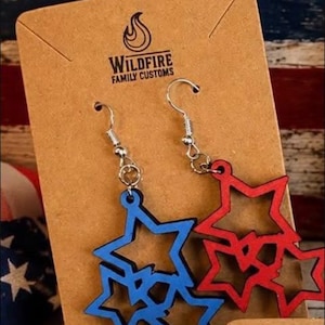 May include: A pair of dangle earrings featuring cutout star shapes attached to silver-toned metal hooks. One earring is blue and the other is red. The earrings are displayed on a brown cardboard card with the text Wildfire Family Customs.