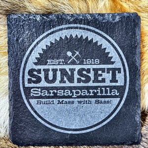 May include: Square black slate coaster with a circular design. The design includes the word "SUNSET" above "Sarsaparilla" and the phrase "Build Mass with Sass!" The coaster also features the text "EST. 1918" and a pickaxe and shovel graphic.