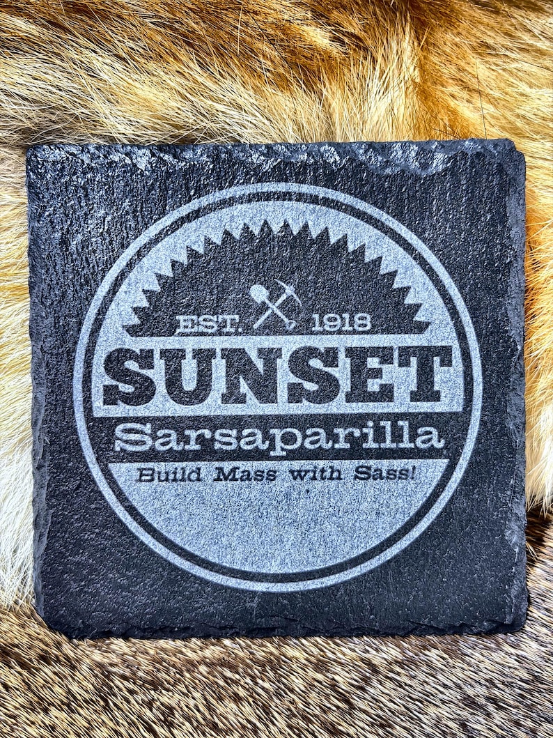 May include: Square black slate coaster with a circular design. The design includes the word "SUNSET" above "Sarsaparilla" and the phrase "Build Mass with Sass!" The coaster also features the text "EST. 1918" and a pickaxe and shovel graphic.