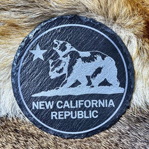 May include: A round, black slate coaster with a white engraved design. The design features two bears, a star, and the text "NEW CALIFORNIA REPUBLIC" in a circular border. The coaster is on a textured, brown and tan surface.