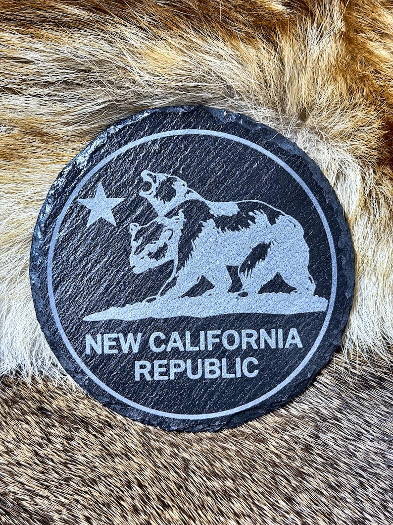 May include: A round, black slate coaster with a white engraved design. The design features two bears, a star, and the text "NEW CALIFORNIA REPUBLIC" in a circular border. The coaster is on a textured, brown and tan surface.