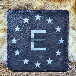 May include: Square black slate coaster with a white letter "E" and surrounding stars. The coaster is made of a dark, textured stone and is designed to protect surfaces from drink condensation.