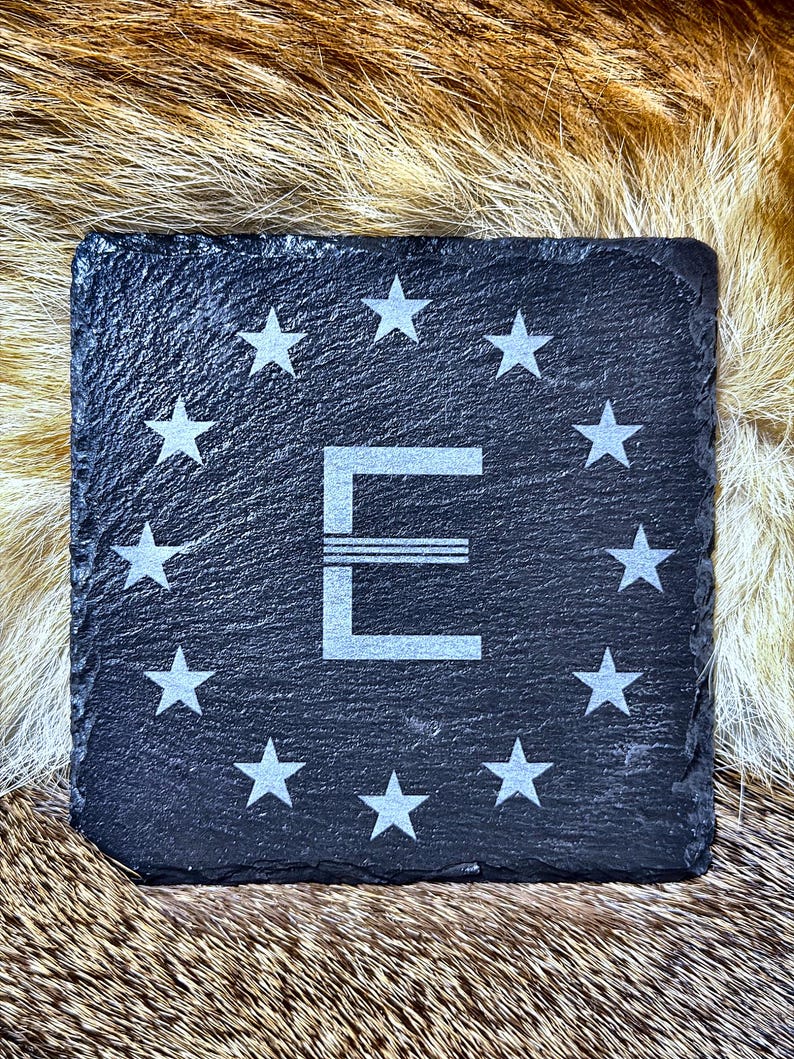May include: Square black slate coaster with a white letter "E" and surrounding stars. The coaster is made of a dark, textured stone and is designed to protect surfaces from drink condensation.