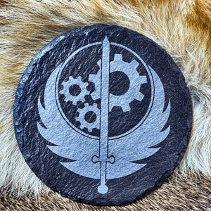 May include: A round, black slate coaster with a gray emblem featuring a sword, gears, and wings. The coaster has a textured surface and is suitable for placing drinks on.