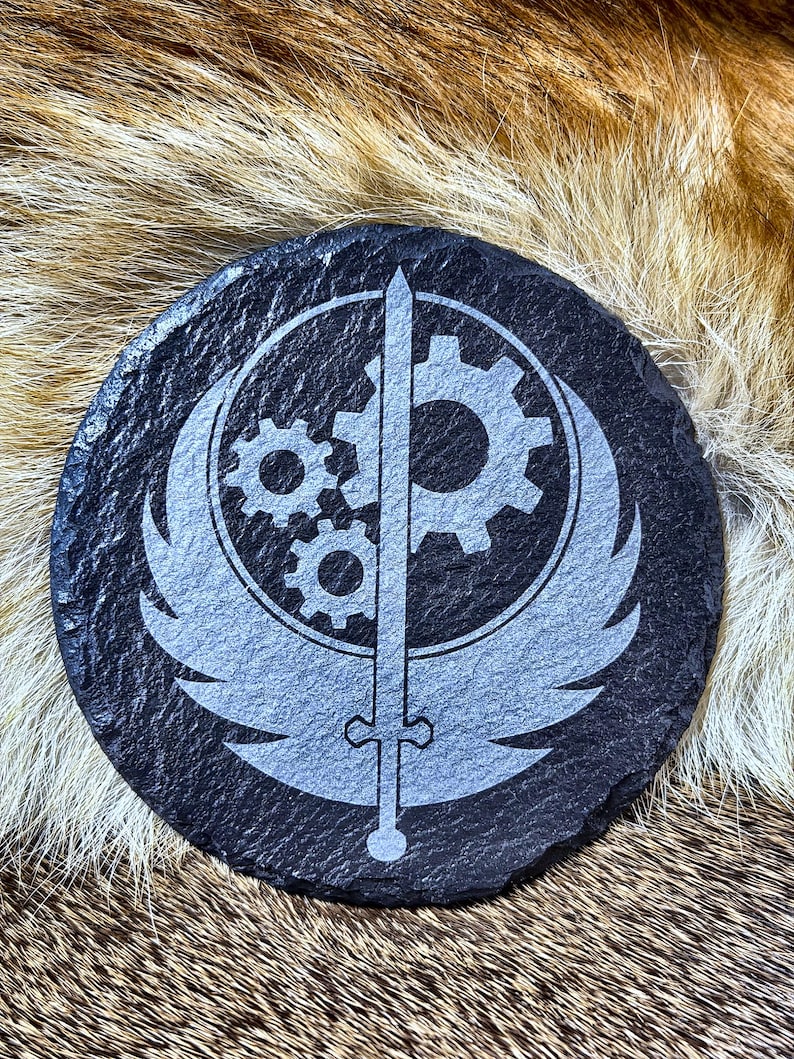 May include: A round, black slate coaster with a gray emblem featuring a sword, gears, and wings. The coaster has a textured surface and is suitable for placing drinks on.