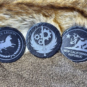 May include: Three round, black slate coasters with etched designs. One features a bull and the text "CAESAR'S LEGION", another has a gear and wing design, and the third displays a bear and the text "NEW CALIFORNIA REPUBLIC".