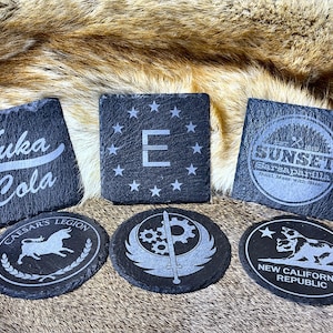 May include: A collection of six slate coasters with etched designs. The coasters feature various logos and text, including "Nuka Cola," "E" surrounded by stars, "Sunset Sarsaparilla," "Caesar's Legion," a gear and sword design, and "New California Republic."