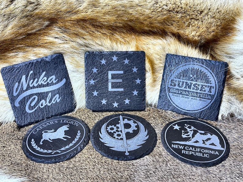 May include: A collection of six slate coasters with etched designs. The coasters feature various logos and text, including "Nuka Cola," "E" surrounded by stars, "Sunset Sarsaparilla," "Caesar's Legion," a gear and sword design, and "New California Republic."