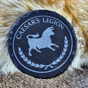 May include: A round, black slate coaster with the words "CAESAR'S LEGION" in a circular border. A silver bull graphic is in the center, surrounded by a laurel wreath design. The coaster is approximately 4 inches in diameter.