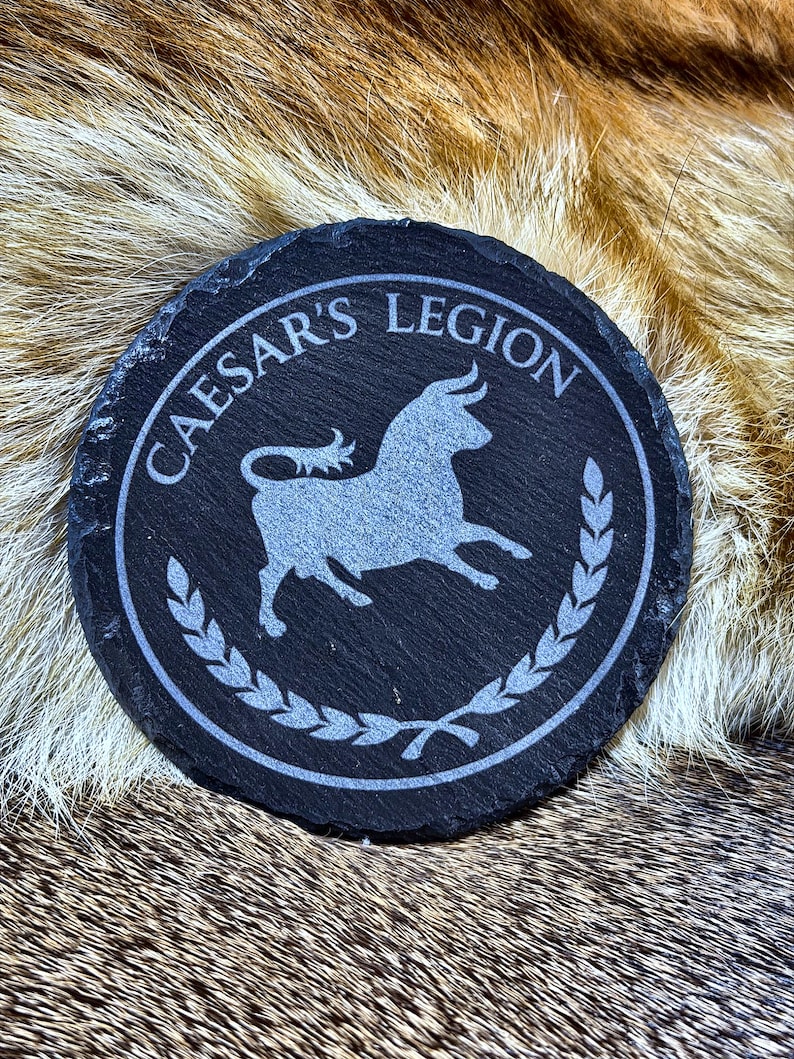 May include: A round, black slate coaster with the words "CAESAR'S LEGION" in a circular border. A silver bull graphic is in the center, surrounded by a laurel wreath design. The coaster is approximately 4 inches in diameter.