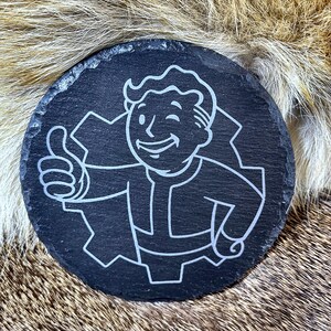 May include: A round, black slate coaster featuring a white graphic of a smiling cartoon figure giving a thumbs-up. The figure is inside a gear-shaped outline. The coaster has a rough, natural edge.