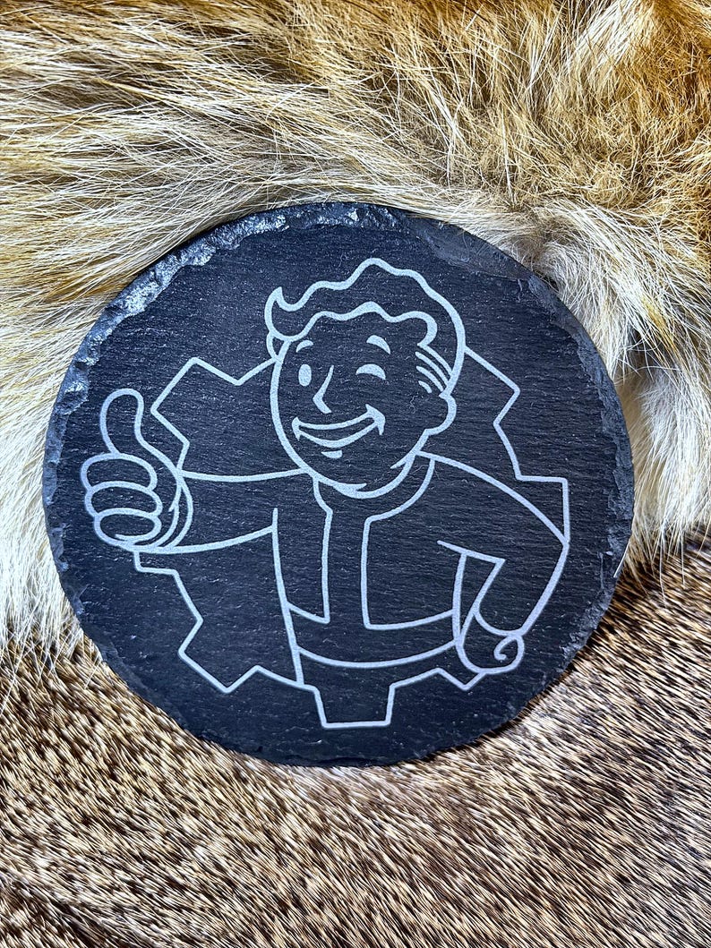 May include: A round, black slate coaster featuring a white graphic of a smiling cartoon figure giving a thumbs-up. The figure is inside a gear-shaped outline. The coaster has a rough, natural edge.