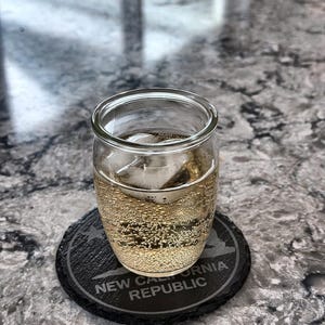 May include: A clear glass tumbler filled with a bubbly, light-colored beverage and ice cubes, resting on a black coaster. The coaster is engraved with the words "NEW CALIFORNIA REPUBLIC" in white.