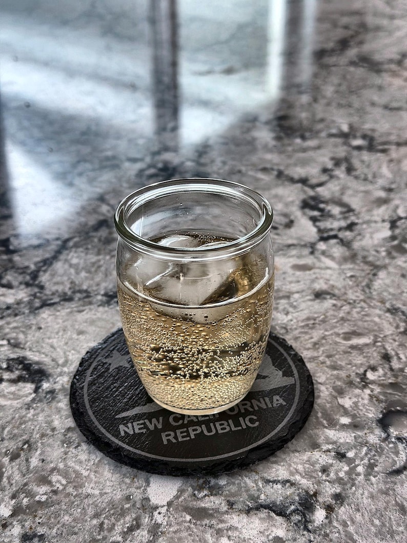 May include: A clear glass tumbler filled with a bubbly, light-colored beverage and ice cubes, resting on a black coaster. The coaster is engraved with the words "NEW CALIFORNIA REPUBLIC" in white.