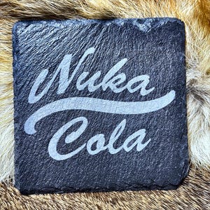 May include: A square, dark gray slate coaster with the words "Nuka Cola" etched in a stylized, light gray font. The coaster has a rough, textured surface and is likely for use with beverages. The overall theme is related to the Fallout video game series.