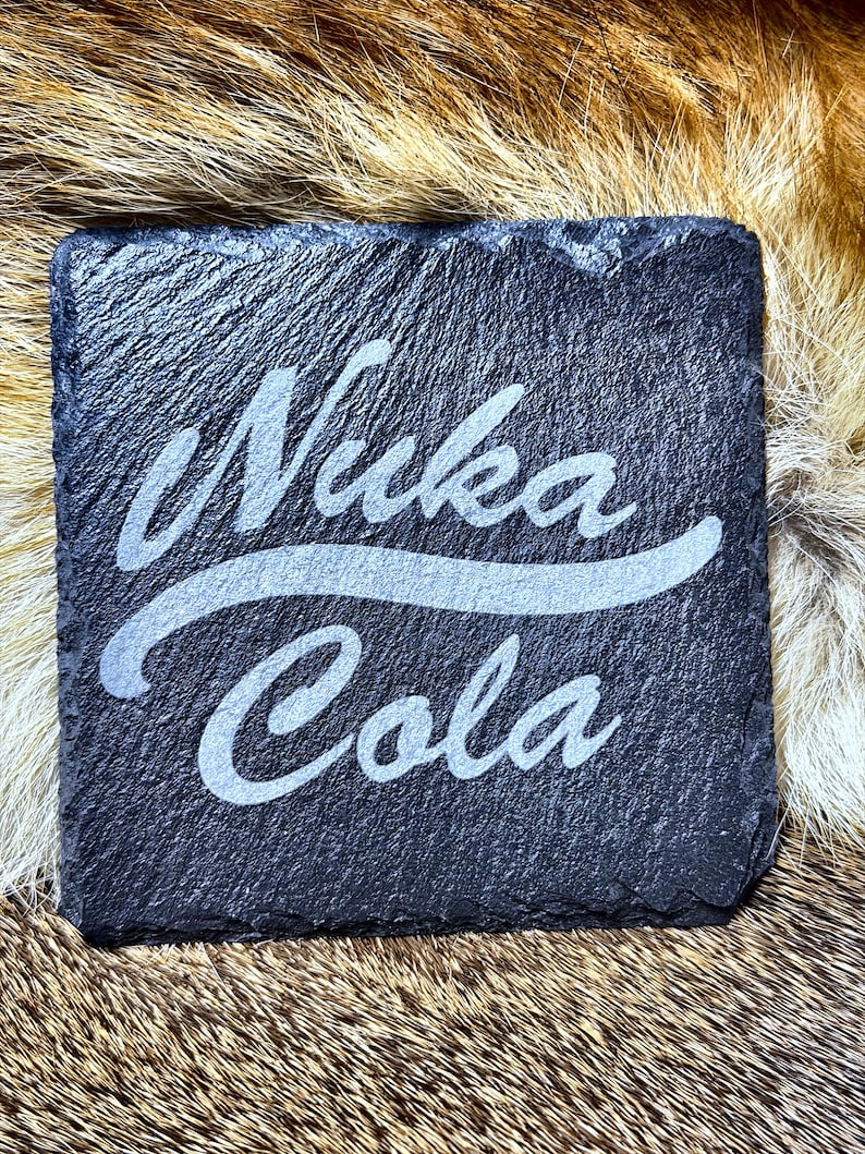 May include: A square, dark gray slate coaster with the words "Nuka Cola" etched in a stylized, light gray font. The coaster has a rough, textured surface and is likely for use with beverages. The overall theme is related to the Fallout video game series.