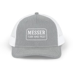 May include: A grey trucker cap with a white mesh back and a curved peak. The front of the cap has a white rectangular patch with the text "MESSER TURF AND PEST". The cap has a snap closure for adjustment.