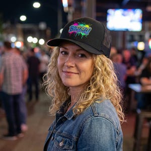 May include: Black trucker hat featuring the word "BOOYAH!" in vibrant, splashy lettering. The hat has a mesh back and a curved peak. The hat is worn by a person with curly blonde hair and a denim jacket.