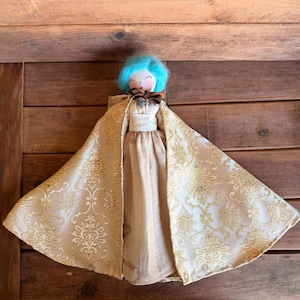 May include: A doll with bright turquoise hair, a painted face with closed eyes, and a brown ribbon tied at the neck. The doll wears a beige dress and a wide, floor-length cape featuring a gold and beige damask pattern with a metallic sheen.