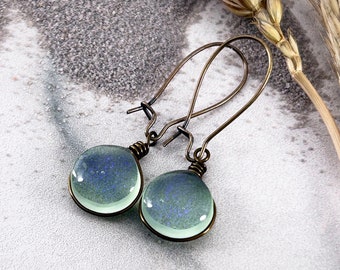 May include: A pair of drop earrings featuring round, translucent light green glass cabochons with subtle blue glitter flecks. The glass pieces are encased in dark bronze-toned wire settings and hang from matching bronze-toned metal ear wires.
