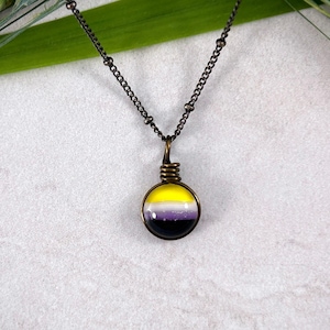 May include: A silver chain necklace with a round pendant featuring the non-binary pride flag colors: yellow, white, purple, and black.