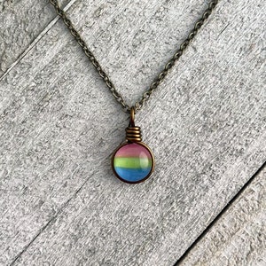May include: A round pendant necklace with a bronze chain. The pendant features a colorful design with pink, green, and blue stripes. The pendant is wire-wrapped in bronze. The necklace is displayed on a gray wooden surface.