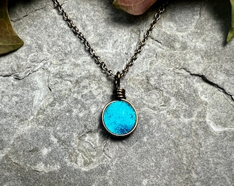 May include: A round pendant necklace featuring a bright turquoise, textured circular center encased in a dark bronze-toned metal wire wrap. The pendant hangs from a matching dark bronze-toned metal chain with small, uniform circular links.