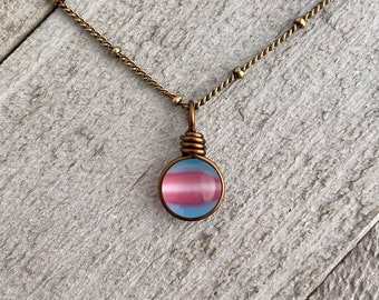 Puede incluir: Un collar de cadena dorada con un colgante redondo que presenta los colores de la bandera del orgullo transgénero en un cabujón de vidrio.