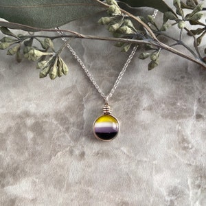 May include: A silver chain necklace featuring a circular pendant with a smooth, domed surface. The pendant displays horizontal stripes in yellow, white, purple, and black, secured by a silver wire wrap at the top connecting to the chain.