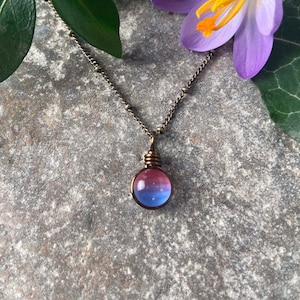 May include: A delicate silver chain necklace with a small, round pendant. The pendant is made of glass and features a gradient of pink, blue, and purple colors.